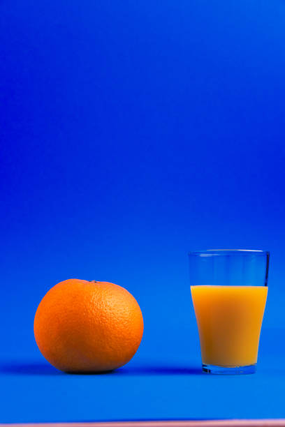 A fresh orange placed next to a glass of orange juice on a vibrant blue background
