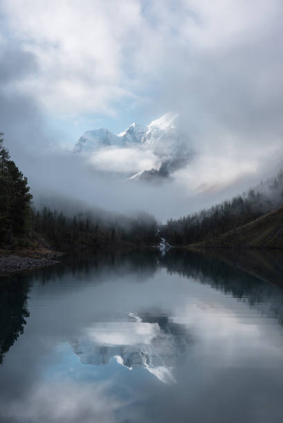 istock Tranquil scenery with snow castle in clouds. Mountain creek flows from forest hills into glacial lake. Snowy mountains in fog clearance, small river and coniferous trees reflected in calm alpine lake. 2170103236