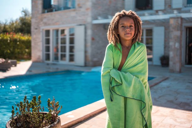 A young mixed race boy standing by the pool, wrapped in a towel.