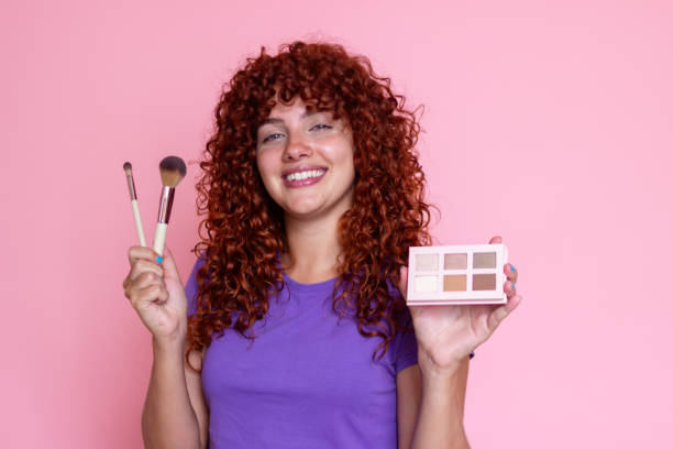 Cheerful redhead woman is holding makeup brushes and an eyeshadow palette in front of a pink background