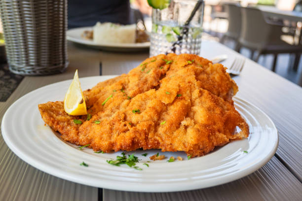 Viennese schnitzel on white plate with glass of drink, second plate on table in outdoor restaurant.