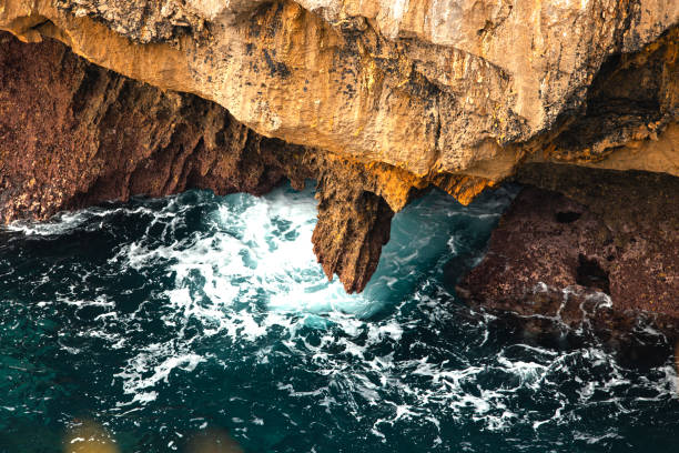 Waves crashing against jagged rocks under rocky overhang, foamy water below .
