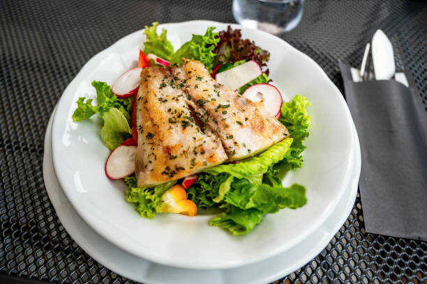 Roasted fish fillet (zander) on fresh green lettuce leaf with radish on white plate, cutlery, closeup.