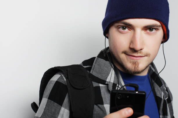 life style, education and people concept: young man listening to music and using smartphone, while standing against grey background.