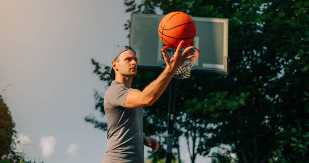 istock A man in a cap confidently spins a basketball on his finger with a focused expression, standing in front of an outdoor hoop. The natural light and greenery create a dynamic, action-filled scene. 2170184477
