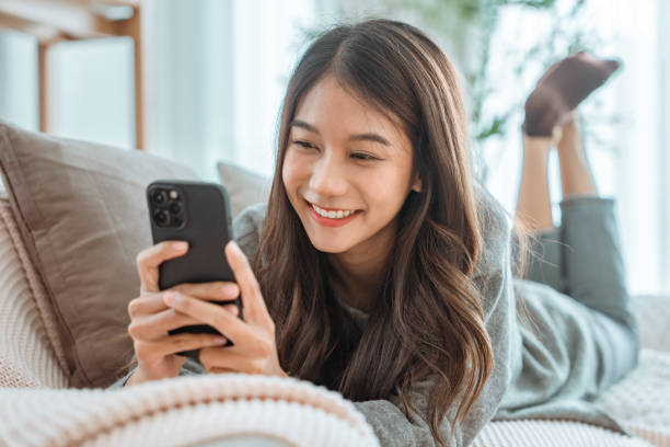 istock Happy young asian woman lady browsing surfing wireless internet on mobile phone while sitting a sofa couch in living room at home apartment, Shopping online via website 2170199633
