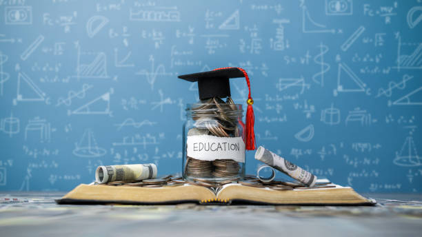 istock concept Student Success Scholarships, Students for College and University Tuition, Saving Money, Tuition for Education, Investment and Scholarships. Coins in a glass jar placed on a book with a graduation cap 2170213197