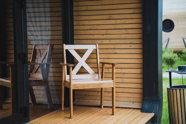The foreground features a weathered, light brownish-pink wooden chair with curved armrests. The chair and its seat appear slightly worn and distressed, showing the wood grain and signs of age.  A simple, matching wooden table is placed in front of it. Both the chair and the table are placed on a wooden floor made of long planks or beams oriented in a horizontal direction. The wood of the floor is a reddish-brown color.
