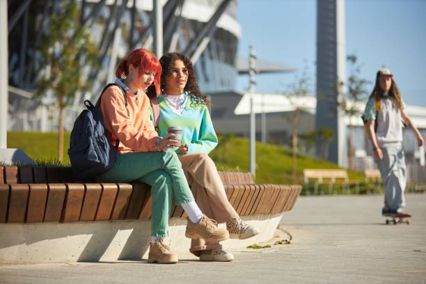 Two teenage girls of diverse ethnicity dressed in bright vibrant clothes sitting together on wooden park bench during break enjoying sunny day on college campus, copy space