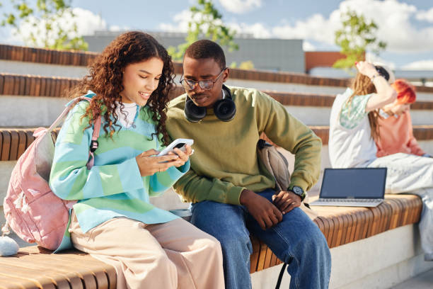 istock Smiling Girl and Male Friend Using Phone Outdoors on Bench 2170231966