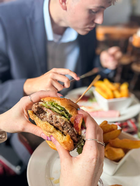Stock photo showing close-up view of a bacon cheeseburger served in a bread bun with beef patty and bacon rasher, lettuce leaves, tomato and melting cheese being held together by wood skewer in order to be eaten by an unrecognisable woman in a pub restaurant setting. In the background a smartly dressed, young man can be seen eating a meal of ham slices, fries in a white bowl, a fried egg and a small side salad of lettuce, red onion and tomato.\n\n* Not AI modified *