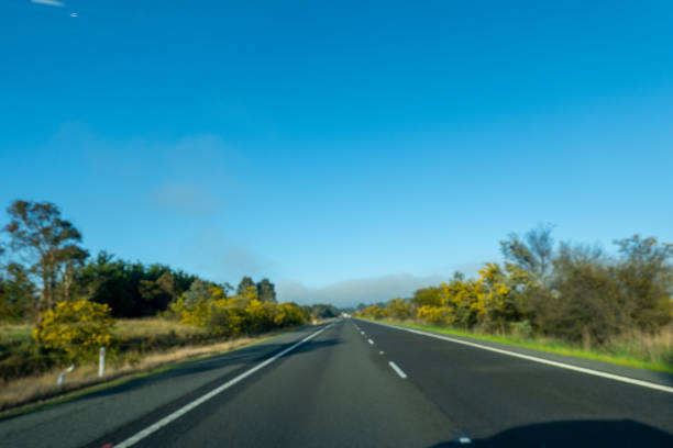 The view from inside a car cruising down a motorway on a clear day, with a bright blue sky stretching across the horizon. The road ahead is clearly visible, with well-defined lane markers and road signs guiding the way. Vehicles in the distance move smoothly along the asphalt, and the surrounding landscape is vibrant under the sunlight.