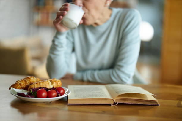 Close up of book and breakfast with unrecognizable woman drinking milk at home.