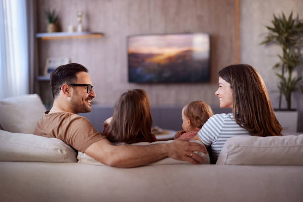 Back view of happy family watching TV on sofa at home. Rear view of happy parents talking while watching TV with their small girls in the living room. couch back stock pictures, royalty-free photos & images