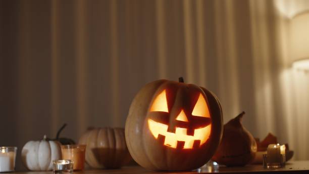 istock Pumpkins on the Table, with a Large Jack-o'-Lantern at the Center with a Flame Inside. Curtains Blowing in the Wind in the Background, Creating a Spooky Home Setting. Background. 2170299186