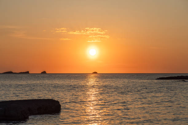 A captivating sunset at Cala Conta beach in Ibiza, Spain, features the sun setting over calm waters with a golden glow. Silhouetted islands dot the horizon, and the tranquil sea reflects the vibrant sky colors