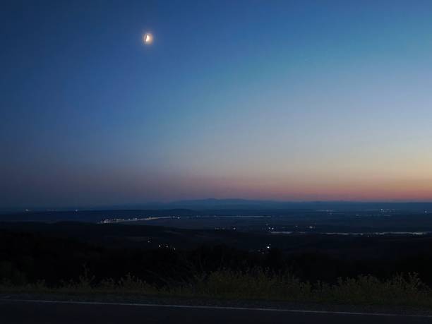 A serene twilight landscape with a crescent moon over a distant horizon and a calm sky transitioning from day to night.