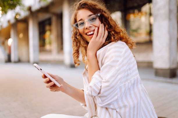istock Young woman enjoying a sunny day while using her phone in a vibrant urban setting with stylish sunglasses and a cheerful expression. 2170310767