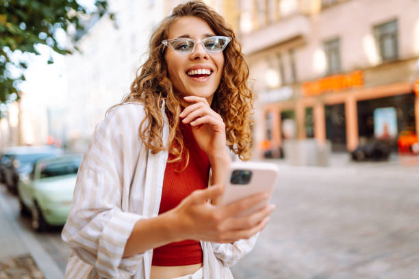 istock Young woman enjoying a sunny day while using her phone in a vibrant urban setting with stylish sunglasses and a cheerful expression. 2170310771