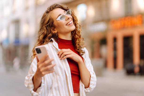 istock Young woman enjoying a sunny day while using her phone in a vibrant urban setting with stylish sunglasses and a cheerful expression. 2170310775