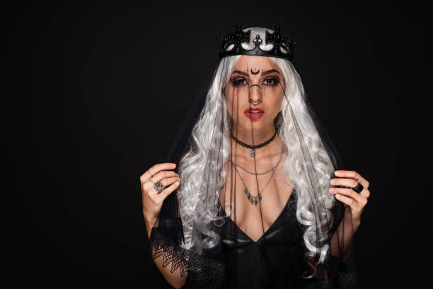 A young woman in a dramatic Halloween costume showcases her intricate wig and striking accessories in a dark studio.