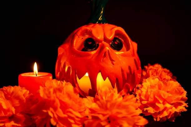 A horizontal photo of a hollowed-out Halloween pumpkin staring forward, lit from within by candles, with a red candle and orange marigolds beside it on a black background