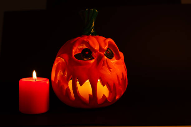 A hollow carved pumpkin with candles inside glows warmly in this horizontal photo. A red candle is lit on the left side, with a deep black background creating contrast