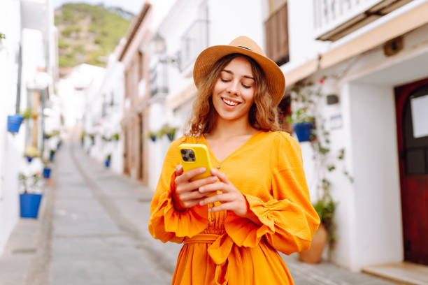 istock A young traveler woman in an orange dress enjoys using her smartphone while strolling through a charming whitewashed street in a sunny village. 2170321026