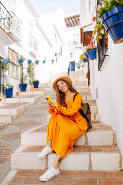 istock A young traveler woman in an orange dress enjoys using her smartphone while strolling through a charming whitewashed street in a sunny village. 2170321028