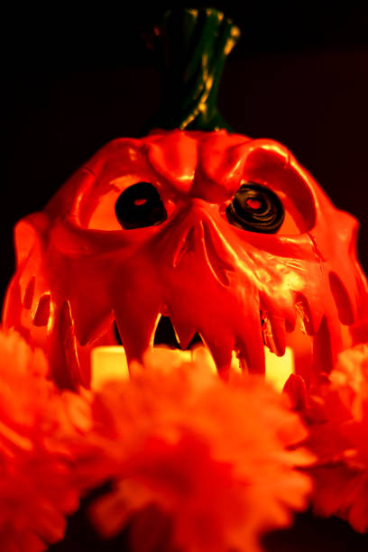 A vertical photo of a hollowed-out Halloween pumpkin with glowing candles inside. A red lit candle and orange marigolds sit beside it, all against a black background
