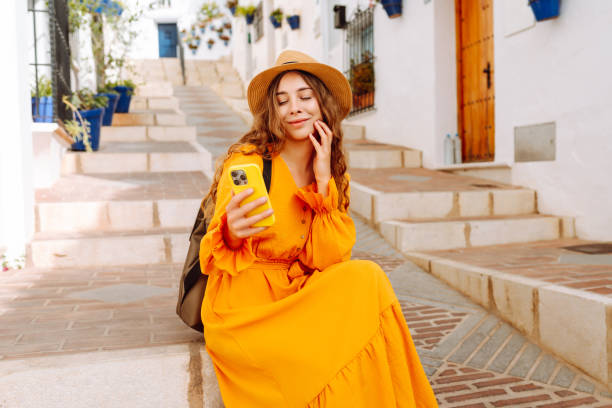 istock A young traveler woman in an orange dress enjoys using her smartphone while strolling through a charming whitewashed street in a sunny village. 2170321076