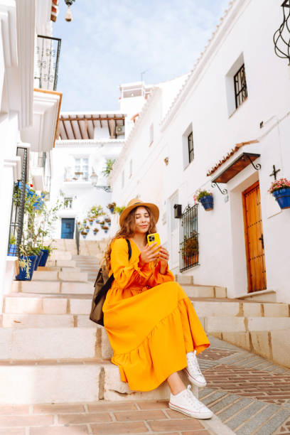 A young traveler woman in an orange dress enjoys using her smartphone while strolling through a charming whitewashed street in a sunny village.