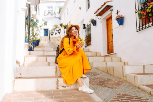 istock A young traveler woman in an orange dress enjoys using her smartphone while strolling through a charming whitewashed street in a sunny village. 2170321093