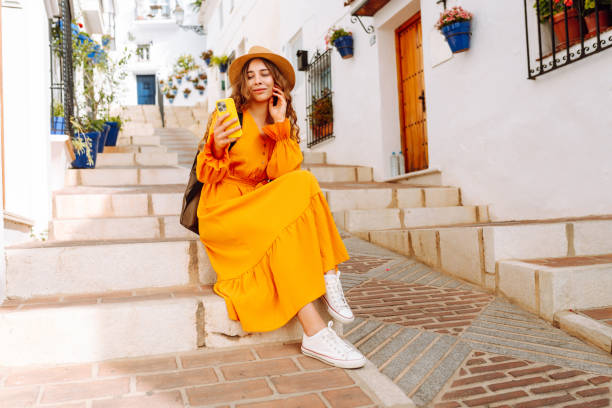 istock A young traveler woman in an orange dress enjoys using her smartphone while strolling through a charming whitewashed street in a sunny village. 2170321114