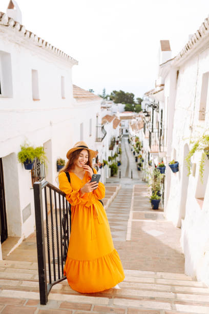 istock A young traveler woman in an orange dress enjoys using her smartphone while strolling through a charming whitewashed street in a sunny village. 2170321134