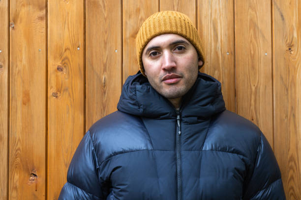 Head and shoulders portrait of a mid-adult male standing in front of a wooden structure whilst staying at luxury glamping pods at a Blossom Plantation in Northumberland, North East England. He is looking directly at the camera with a neutral expression, enjoying a peaceful moment of solitude in nature. He is wearing warm clothing on a cool spring day.\n\nVideos similar to this scenario available.