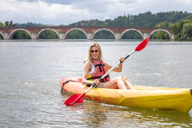 Medium shot of a woman kayaking down the River Tarn in Moissac, France. She is wearing casual clothing with a red life vest over the top. The kayak is yellow and red with a red oar. The woman is wearing sunglasses while smiling.\n\nVideos are available similar to this scenario.
