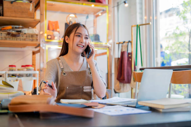 istock leather shop leather bag designer talking to customer smartphone checking order with laptop business female owner working in home studio handcraft product 2170380021