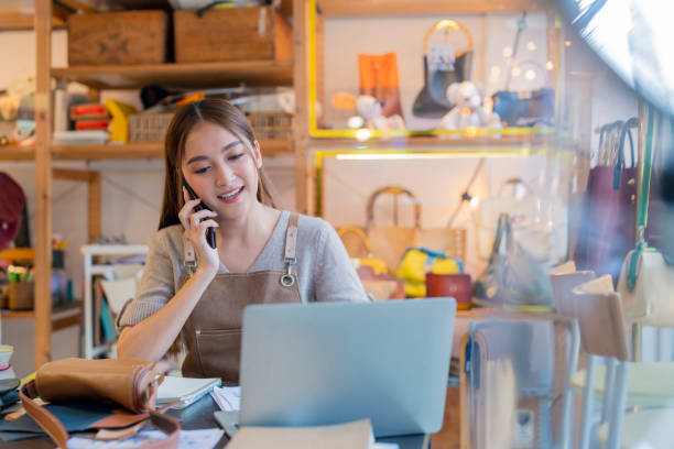 istock leather shop leather bag designer talking to customer smartphone checking order with laptop business female owner working in home studio handcraft product 2170380064