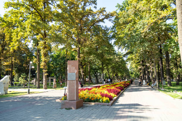 Bishkek, Kyrgyzstan - August 31, 2024: A scenic walkway in Oak Park lined with trees, a monument, and colorful flower beds on a bright day with people strolling