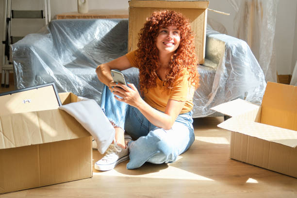 istock Woman sitting on floor using smartphone surrounded by cardboard boxes 2170387596