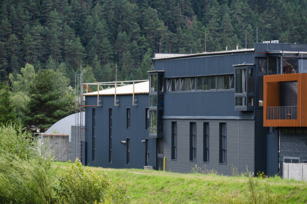 A modern industrial building with a dark facade is set against a lush, forested background. The building's sleek design features large windows and metal siding, blending contemporary architecture with the natural surroundings.