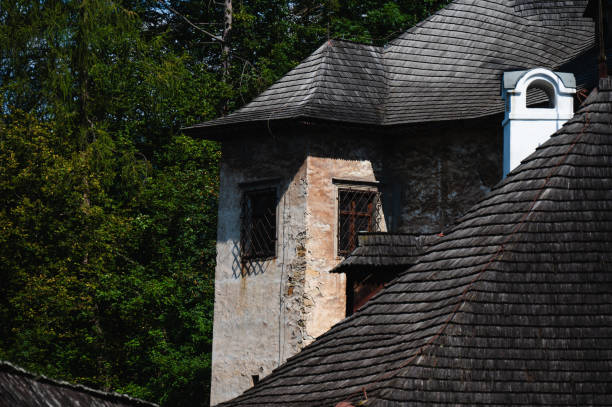 An old stone tower with small windows and a wooden shingle roof is nestled in a forest setting. The weathered walls and traditional roof design reflect the building's historical character, surrounded by lush green trees.