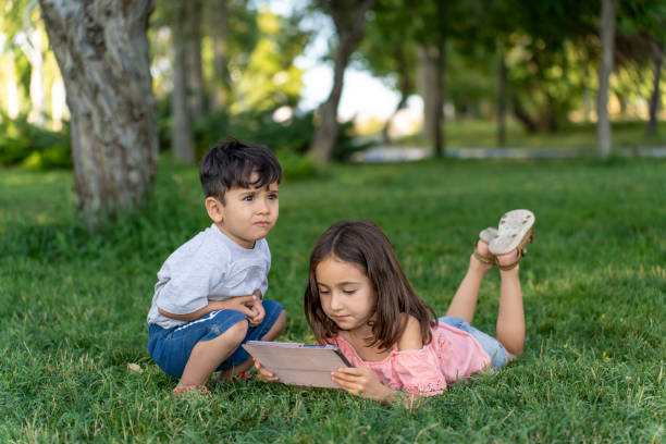 Boy and girl lying on grass using tablet.  Boy and girl watching video on tablet outdoors. Primary school students on summer vacation.