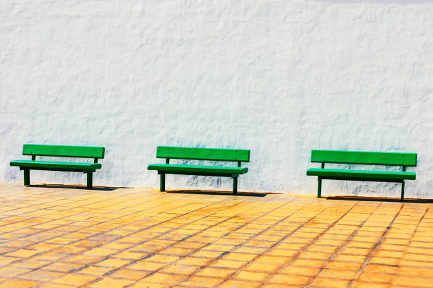 Green benches are sitting on a sidewalk in front of a white wall. Wooden benches are empty and the area is quiet