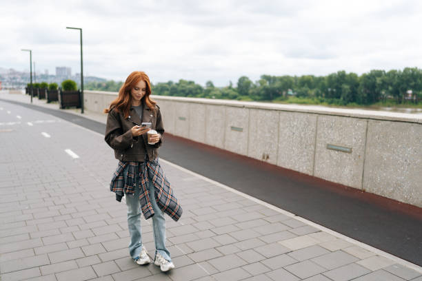 istock Remote view of pretty young woman happily strolling by riverfront, enjoying coffee break while checking phone on cloudy day. 2170436162
