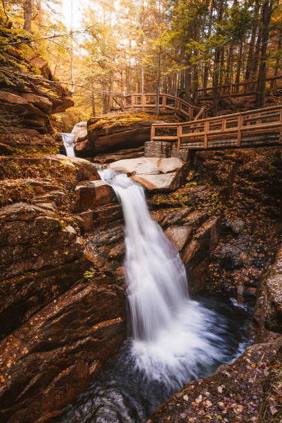 istock Beautiful Sabbaday Falls New Hampshire White Mountain National Forest. Smooth Waterfall Cascading Rocky Edge Forest Trail Wooden Bridge Sun Flare 2170441034