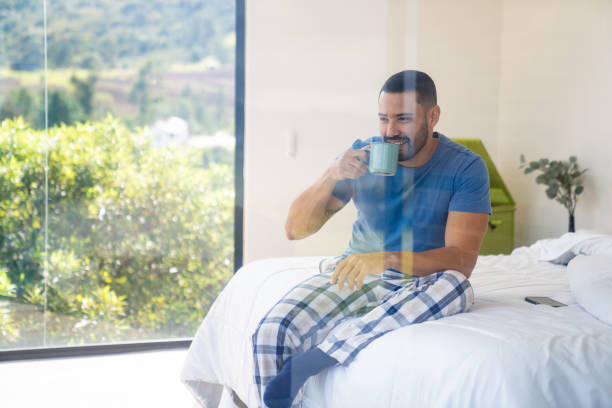 Happy Latin American man in his bedroom drinking a cup of coffee -photographed through window