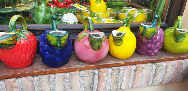 A row of handcrafted ceramic jugs is displayed on a wooden shelf against a white textured wall in bright sunlight. Each pitcher features hand-painted floral motifs and traditional architectural designs, accompanied by colorful knitted and crocheted textiles in the background. The scene captures the charm of artisan craftsmanship and the vibrant atmosphere of a local outdoor market.