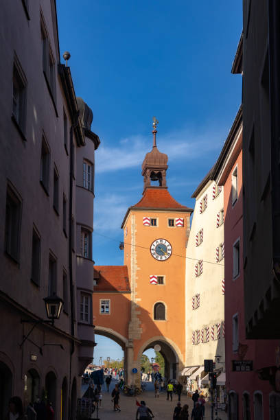 A picturesque view of a historic clock tower in Regensburg, Bavaria, featuring charming architecture and bustling street life on a sunny day.